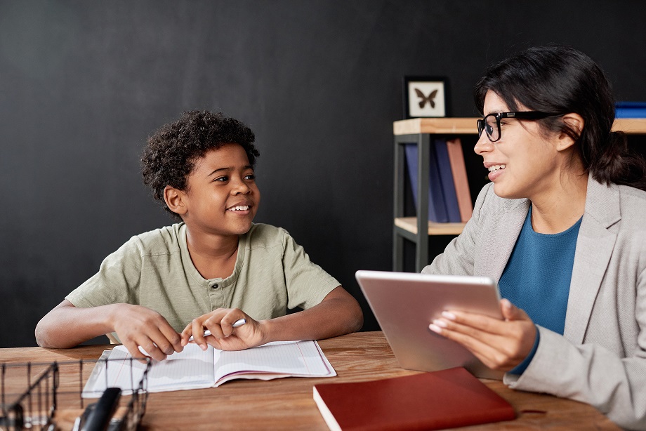 Smiling Latin teacher in glasses sitting at table and using tablet while assisting boy with homework at tutorial